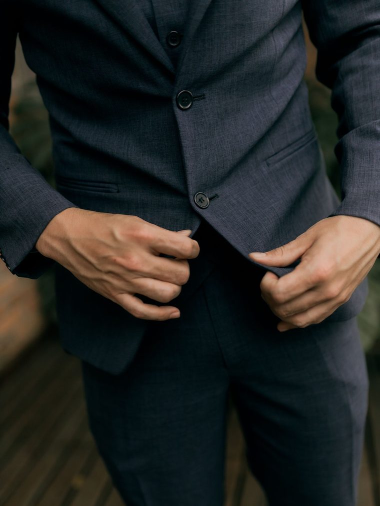 A stock photograph, not a suit we made. We'll replace it with our own once the first suits are finished. A man fastening the button of a navy tailored suit jacket.
